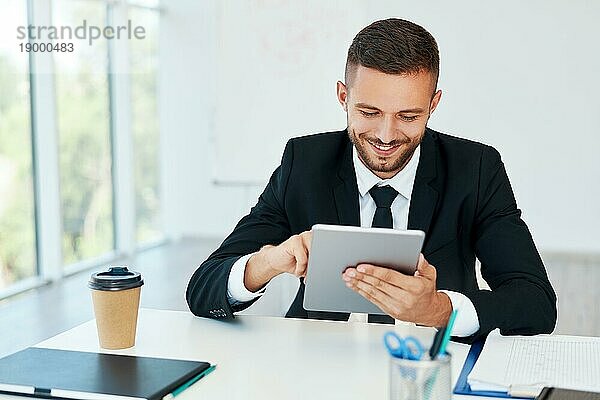 Glücklicher erfolgreicher Geschäftsmann in eleganter Suite mit Tablet in modernem kreativen Büro sitzend. Geschäftskonzept