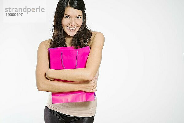 Begeisterte junge Frau  die ihre Einkäufe in einer hübschen lila Einkaufstasche besitzergreifend an ihre Brust drückt  während sie glücklich lächelt und ihre Zufriedenheit zeigt  vor weißem Hintergrund
