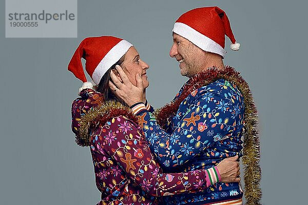 Festliche romantische Paar feiert Weihnachten in Santa Hats und bunten Xmas themed Kleidung umarmt einander mit glücklichen Lächeln in einer Seitenansicht vor einem grauen Hintergrund