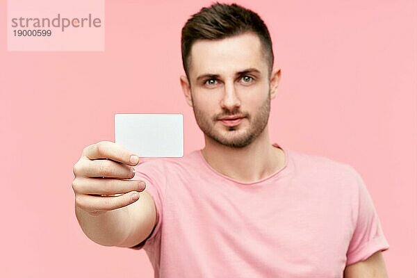 Porträt eines jungen Mannes  der seine Kreditkarte zeigt und in die Kamera schaut  vor einem rosa Hintergrund