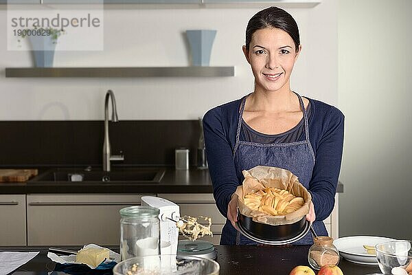Lächelnde  attraktive junge Frau beim Backen  die in ihrer Küche steht und stolz ihre frische Apfeltorte auf einer mit frischen Äpfeln und den Resten der Zutaten bestückten Theke präsentiert