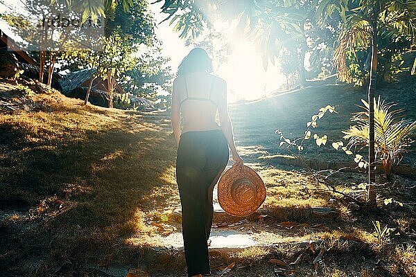 Rückenansicht einer jungen Frau mit Strohhut  die eine Treppe in einem grünen tropischen Park im Sonnenlicht hinaufgeht. Erfolg  Ziel Konzept
