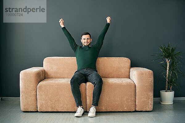Fröhlicher junger Mann mit erhobenen Armen  der seinen Erfolg auf dem Sofa in einer modernen Wohnung feiert. Gewinnerkonzept
