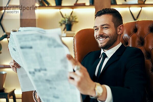 Glücklicher junger Geschäftsmann liest Zeitung und die neuesten Nachrichten  während er morgens in einem Sessel in einem modernen Interieur sitzt