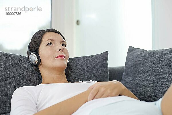 Schöne  ruhige junge Frau  die sich mit einem sinnlichen Glitzern in den Augen entspannt und Musik über Kopfhörer hört  auf dem Rücken liegend auf einem Sofa in lässigen Shorts