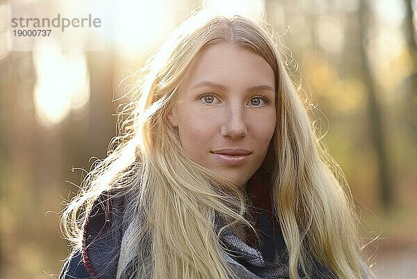 Attraktive junge Frau mit langen blonden Haaren in Herbstmode  die einen trendigen Schal trägt  steht im Freien in einem herbstlichen Wald und lächelt in die Kamera  Kopierraum zur Seite