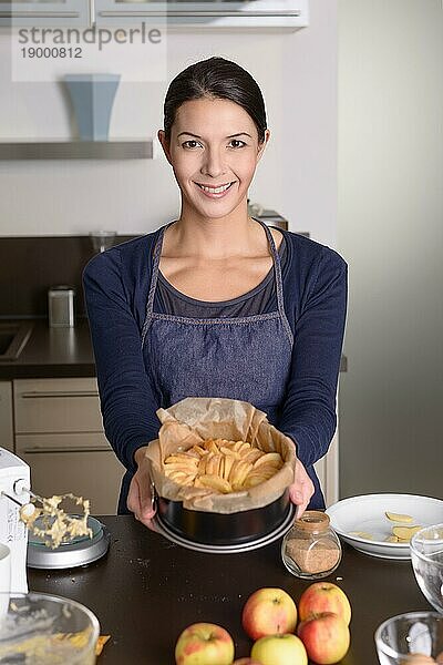Lächelnde  attraktive junge Frau beim Backen  die in ihrer Küche steht und stolz ihre frische Apfeltorte auf einer mit frischen Äpfeln und den Resten der Zutaten bestückten Theke präsentiert