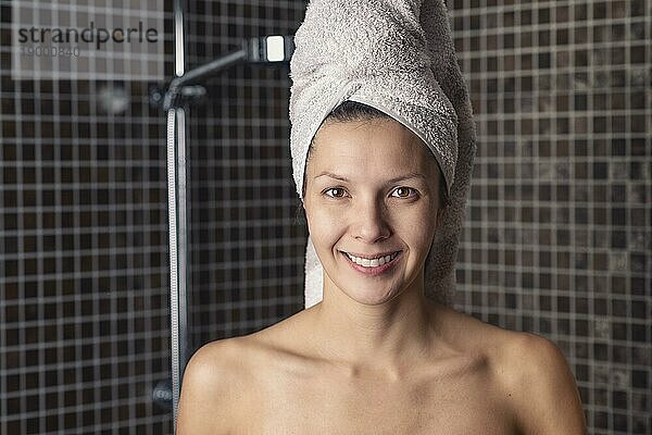 Glückliche nackte attraktive Frau mit ihren naßen Haaren in einem Handtuch  die in ihrem Badezimmer steht und in die Kamera lächelt  in einem Gesundheits  Schönheits und Hygienekonzept