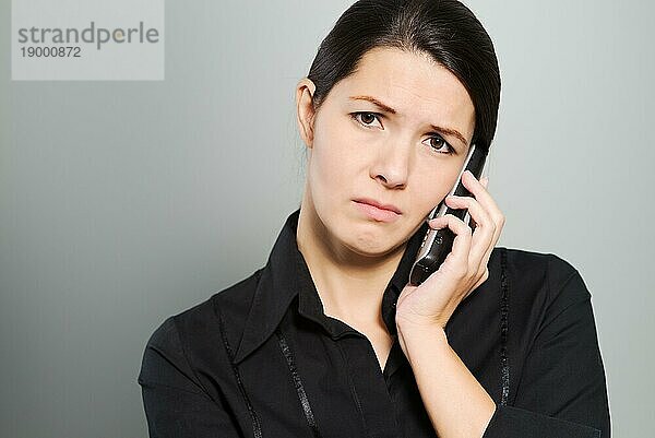 Attraktive Frau  die mit einem traurigen Gesichtsausdruck einem Gespräch über ihr Handy oder Smartphone zuhört  während sie in die Kamera schaut