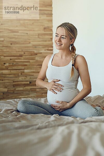 Glückliche junge schwangere Frau  die mit ihrem ungeborenen Kind auf einem Bett sitzt und ihren Babybauch mit den Händen wiegt und mit einem zärtlichen Lächeln der Liebe zur Seite schaut