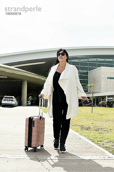 Porträt einer reifen Geschäftsfrau in eleganter Kleidung  die einen Koffer auf Rädern trägt und den Flughafen verlässt. Geschäft  erfolgreiche Menschen  Transportkonzept