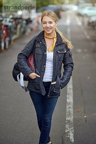 Porträt einer jungen blonden Frau auf der Straße in der Stadt  die ihre Hände in ihrer lässigen schwarzen Jacke ohne Reißverschluss hält  in die Kamera schaut und lächelt