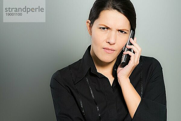 Nervöse  hübsche junge Frau  die mit ihrem Handy telefoniert und mit einem verwirrten Gesichtsausdruck und Stirnrunzeln in die Kamera schaut  vor einem grauen Studiohintergrund