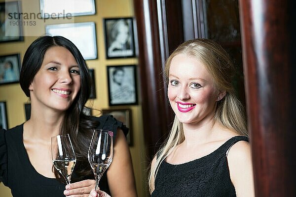 Zwei schöne  elegante Frauen in stilvollen  schlichten schwarzen Cocktailkleidern trinken ein Glas gekühlten Champagner  während sie einen gemeinsamen Abend feiern