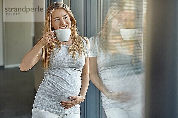 Glückliche  gesunde  schwangere  junge  blonde Frau  die an ein Terrassenfenster gelehnt steht und in die Kamera lächelt  während sie eine Tasse Tee genießt