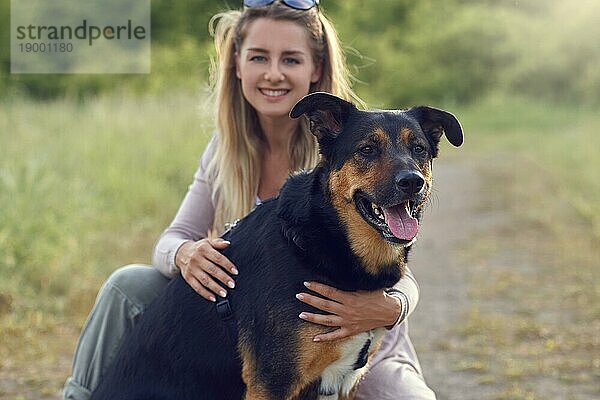 Hübsche blonde Frau mit ihren beiden Hunden  die am Rande einer Landstraße mit langem Frühlingsgras kauern und sich auf einen großen braunen Hund im Vordergrund konzentrieren