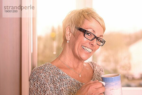 Lebhafte attraktive blonde Frau mittleren Alters mit Brille  die Kaffee trinkt und mit einem strahlenden Lächeln zur Seite in die Kamera blickt