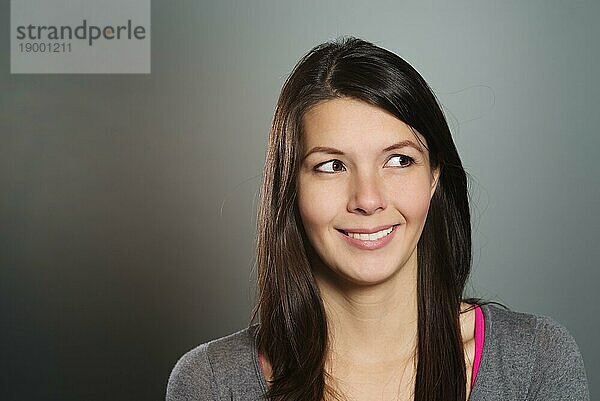 Attraktive junge Frau mit schönem  freundlichem Lächeln  die mit abgewandtem Blick seitlich nach links blickt  Kopf Schulter Studioporträt auf Grau