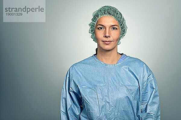 Attraktive Ärztin oder Krankenschwester mittleren Alters in Kittel und Sterilitätskappe  die vor einem grauen Hintergrund direkt in die Kamera schaut  Kopf Schulter Porträt