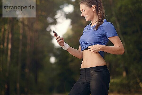 Junge  sportliche Frau  die im Wald trainiert und auf ihr Smartphone schaut  während sie eine mobile Anwendung nutzt  die elektronisch mit ihrem Pulsgurt verbunden ist