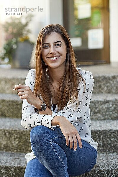 Attraktive glückliche junge Frau mit langen braunen Haaren  die Jeans trägt und entspannt auf einer Stufe im Freien sitzt und in die Kamera schaut  mit einem schönen warmen strahlenden Lächeln