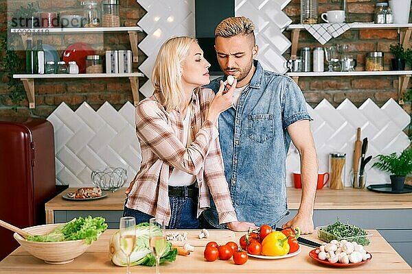 Junger Mann weigert sich  Gemüse zu essen. Frau füttert Mann mit gesunden Lebensmitteln beim Kochen in der Küche zu Hause. vegetarisches Konzept