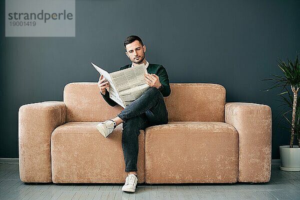 Selbstbewusster junger Mann liest Zeitung und die neuesten Nachrichten  während er morgens auf dem Sofa in einem modernen Interieur sitzt