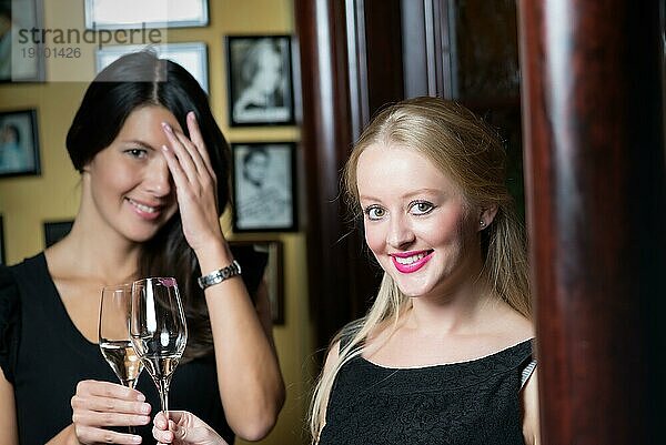 Zwei schöne  elegante Frauen in stilvollen  schlichten schwarzen Cocktailkleidern trinken ein Glas gekühlten Champagner  während sie einen gemeinsamen Abend feiern