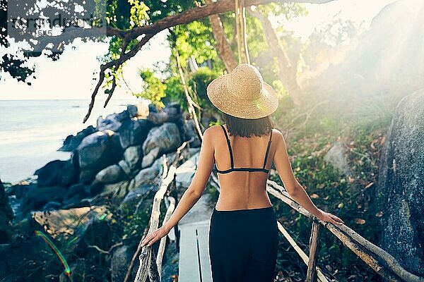 Rückansicht einer jungen Frau mit Strohhut  die auf einer Holzbrücke auf einer tropischen Insel im Gegenlicht spazieren geht. Urlaub  Reisekonzept