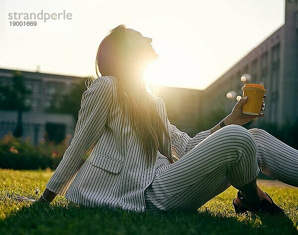Junge stilvolle Frau trinken Kaffee und entspannen Sie sich auf Stadtpark Gras im Sonnenschein Licht sitzen. Pause Zeit Konzept