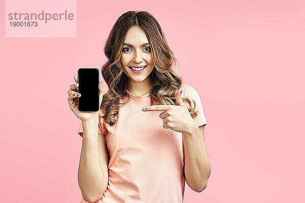 Lächelnde hübsche Frau  die mit dem Finger auf ihr Handy zeigt und in die Kamera schaut  auf rosa Studiohintergrund