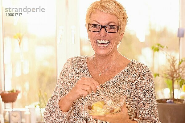 Lebhafte attraktive Frau mittleren Alters mit Brille  die eine gesunde Schüssel vitaminreichen Obstsalat ißt und mit einem freundlichen Lächeln voller Lebensfreude in die Kamera schaut