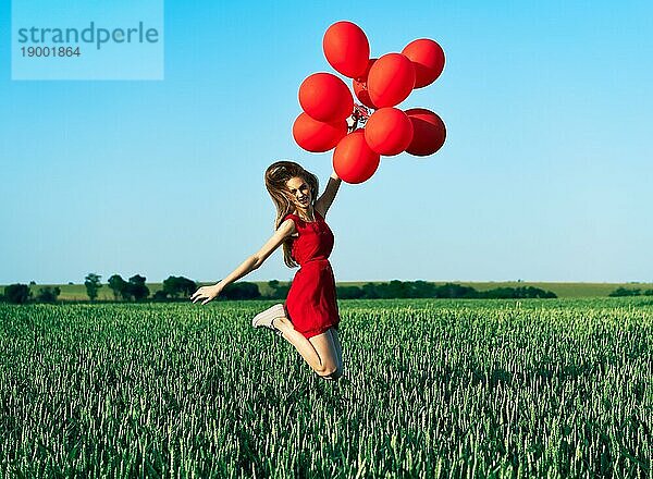 Junge glückliche Frau springt mit roten Luftballons auf grünem Sommerfeld. Spaß  Glück  Freiheit  Feier Konzept