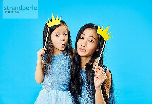 Mutter und niedliche kleine Tochter mit einem Papier Krone auf Stick auf blauem Hintergrund haben Spaß zusammen. Glücklich lustige Familie Konzept