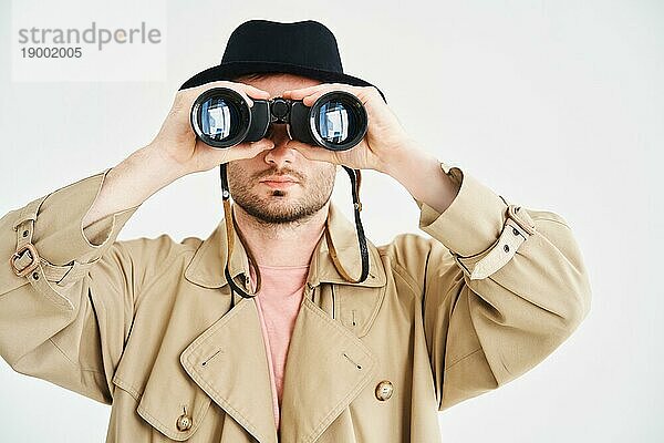 Junger Mann in Mantel und Hut schaut durch ein Fernglas auf isoliertem weißen Hintergrund. Spion  Spionage Konzept