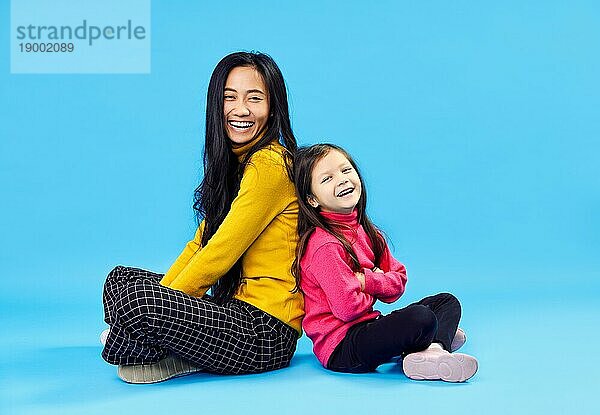 Glückliche lächelnde Mutter und ihre kleine süße Tochter posieren auf blauem Studio Hintergrund