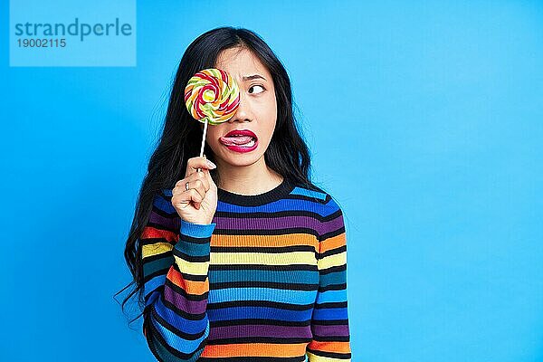 Close up von jungen asiatischen Frau streckt die Zunge zu bunten Lutscher vorblauem Hintergrund. Emotionen Konzept  süßes Essen