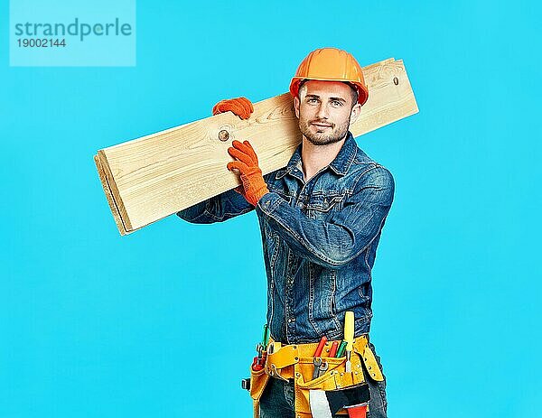Junger gutaussehender Zimmermann hält Holz auf seinen Schultern über blauem Hintergrund und schaut in die Kamera