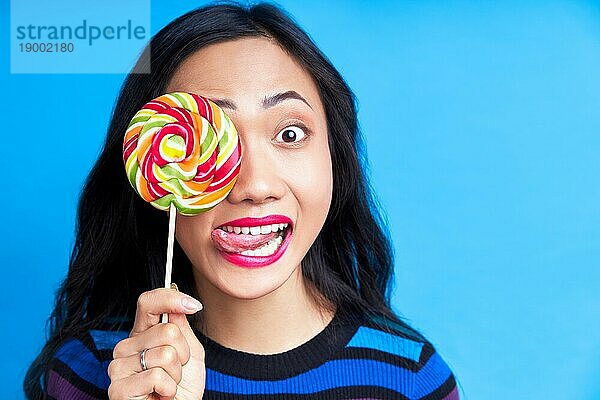 Close up von jungen asiatischen Frau streckt Zunge bedeckt ihr Auge mit bunten Lutscher vorblauem Hintergrund. Emotionen Konzept  süßes Essen