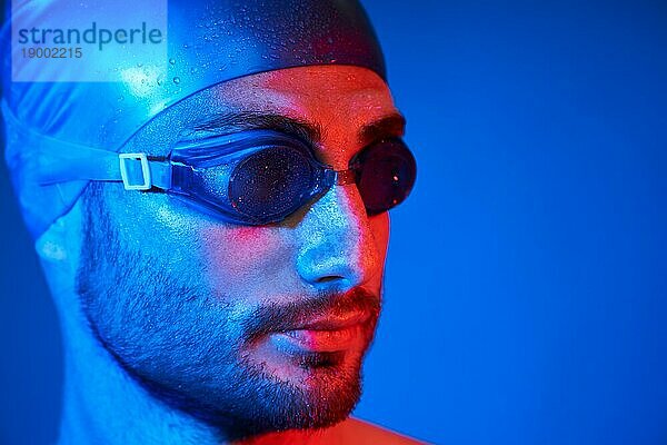Close up Porträt von gut aussehend Schwimmer mit Schwimmbrille in rot rosa Neonlicht über blauem Hintergrund. Sport  Mode  männliche Schönheit