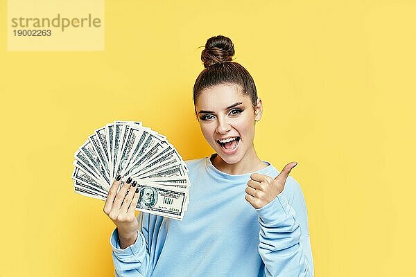 Glückliche aufgeregte junge Frau zeigt Geldscheine mit Daumen nach oben auf gelbem Hintergrund. Motivation  Erfolg Konzept