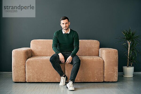 Junger trendiger Mann posiert auf dem Sofa sitzend und schaut in die Kamera. Entspannen Konzept