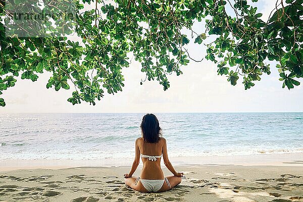 Rückenansicht der jungen Frau meditiert auf tropischen Sandstrand unter Mangrove Baum. entspannen und zen Konzept