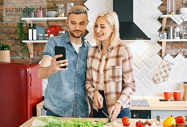 Junges Paar kocht zusammen vegetarische Mahlzeit nach Rezept auf dem Handy in der Küche zu Hause