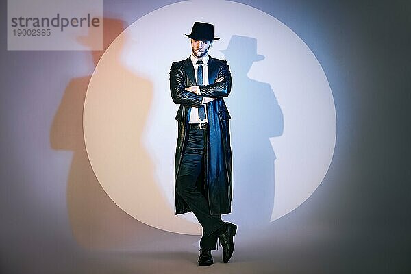 Ganzkörperporträt eines selbstbewussten  gut aussehenden Mannes in schwarzem Mantel und Hut mit verschränkten Armen  der im Studio im Scheinwerferlicht posiert. Noir Film Stil. Privatdetektiv  Spion  Ermittlungskonzept
