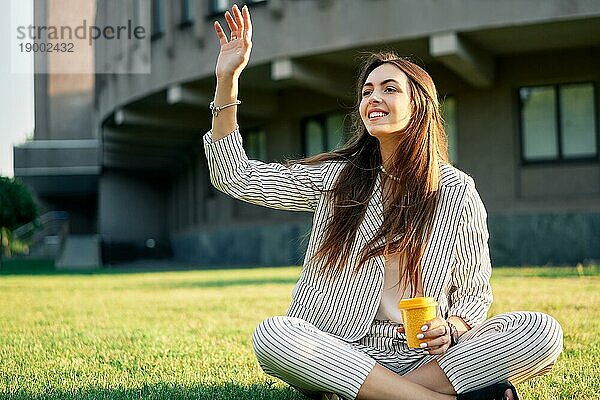 Junge hübsche Frau begrüßen Freunde winken angehoben Palme  sitzen auf Gras mit Kaffee. Hallo  Gruß Konzept