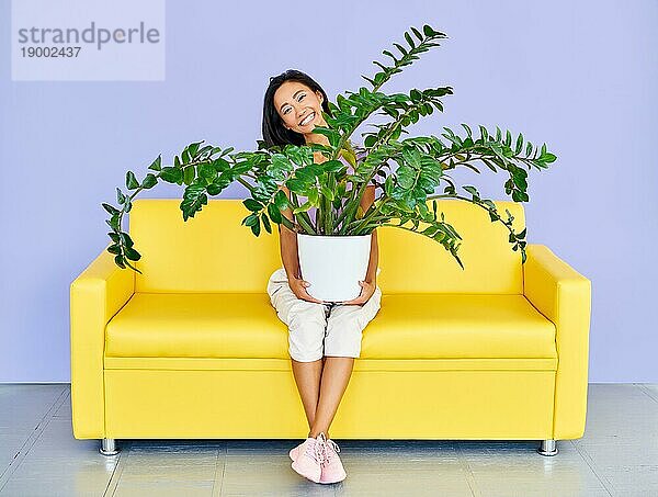 Junge lächelnde Frau sitzt auf dem Sofa und hält große Pflanze im Topf. Spaß  Haus Garten Konzept