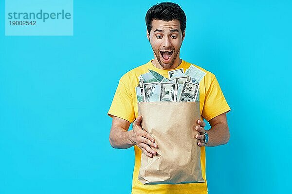 Aufgeregt glücklicher Mann halten Papiertüte voll von Stapeln von Geld vorblauem Hintergrund. Erfolg  Geschäft  und Vermögen Konzept