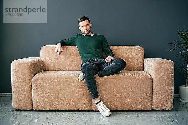 gutaussehender  trendiger Männchen  der zur Kamera schaut  während er auf einem bequemen Sofa in einer modernen Wohnung sitzt. Männliches Porträt