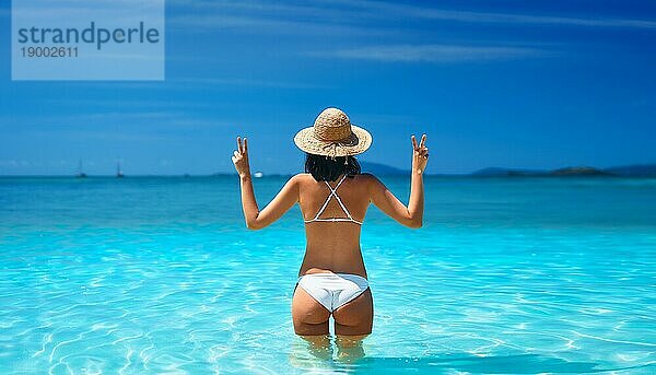 Glückliche sexy Frau in weißem Bikini und Strohhut zeigt Friedenszeichen mit beiden Händen entspannen in türkisfarbenen Meerwasser auf tropischen Strand im Paradies Insel. Entspannen  Urlaub  Sommer Reise Konzept